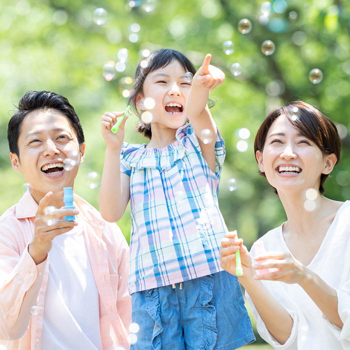 シャボン玉で遊ぶ笑顔の親子三人と、新緑の背景