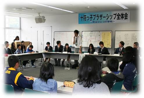 長机をロの字に並べて座っている生徒たちの中、1人の男子生徒が起立し話をしている写真