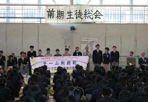 前方に前期生徒総会と書かれた横断幕が壁に貼られ、代表の生徒たちが前に並んで立っているのを生徒たちが床に整列して座っている様子を写した写真
